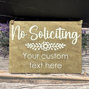 Puede incluir: Un letrero rectangular de madera con las palabras "No Soliciting" en escritura blanca. Debajo del texto hay un diseño floral, y las palabras "Your custom text here" también están en blanco. El letrero tiene una cuerda de yute para colgar.