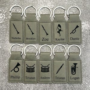 May include: A collection of personalized keychains in a light gray color. Each keychain features a silver metal ring and a rectangular tag with a musical instrument graphic and a name. Instruments include a clarinet, trumpet, drum, and tuba.