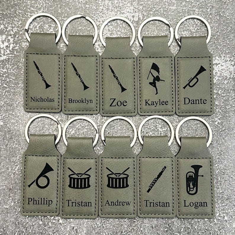 May include: A collection of personalized keychains in a light gray color. Each keychain features a silver metal ring and a rectangular tag with a musical instrument graphic and a name. Instruments include a clarinet, trumpet, drum, and tuba.