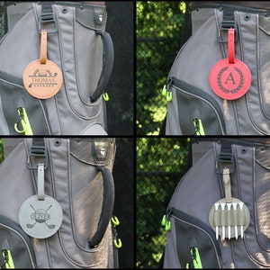 May include: Four golf bag tags in a collage. The tags are round and attached to golf bags. One tag is tan with the name "Thomas" and golf-themed graphics. Another is red with a black "A" and a laurel wreath. The third is gray with "Oliver" and crossed golf clubs. The last is olive green with golf tees.