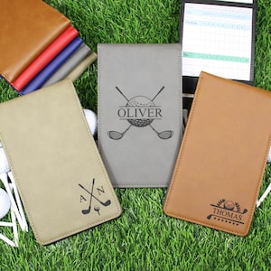 May include: Golf scorecards and covers in various colors, including tan, gray, and brown, are displayed on a bed of green grass. The covers are personalized with names and golf-themed graphics. Golf balls and tees are also visible.