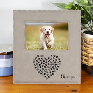 May include: A gray faux leather picture frame with a heart made of paw prints and the word "Champ" inscribed below. The frame has a 4x6 inch photo opening and features a photo of a golden retriever puppy.