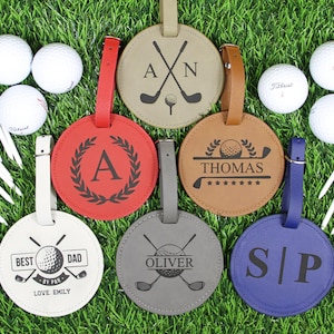 May include: A collection of personalized golf bag tags in various colors, including red, tan, brown, gray, and blue. Each tag features a unique design with initials, names, or golf-themed graphics. Golf balls and tees are also visible.