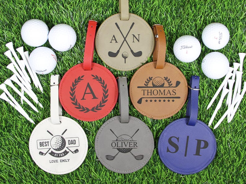 May include: A collection of personalized golf bag tags in various colors, including red, tan, brown, gray, and blue. Each tag features a unique design with initials, names, or golf-themed graphics. Golf balls and tees are also visible.