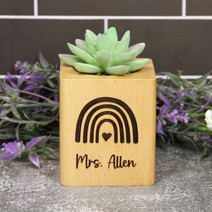 May include: A square light-colored wooden planter containing a green succulent. The front face features a dark brown laser-engraved rainbow graphic with a small heart in the center and the text "Mrs. Allen" written below the rainbow.