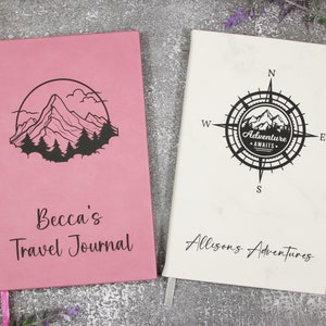 May include: Two pink and white travel journals with mountain and compass designs. The pink journal says "Becca's Travel Journal" and the white journal says "Allison's Adventures".