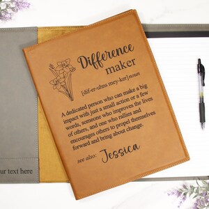 May include: A brown leather journal with a gray leather flap. The journal has a floral design and the text "Difference maker [dif-er-uhns mey-ker] noun A dedicated person who can make a big impact with just a small action or a few words, someone who improves the lives of others, and one who rallies and encourages others to propel themselves forward and bring about change. see also: Jessica" engraved on the front.