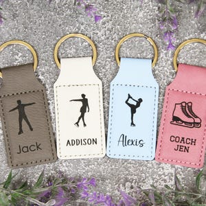 May include: Four keychains with gold rings. Each keychain has a different color: gray, white, light blue, and pink. Each keychain has a black graphic of a figure skater and a name or title: Jack, Addison, Alexis, and Coach Jen. The keychains are for ice skating.
