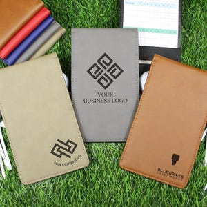 May include: Three leather golf score card holders in tan, gray, and brown, with golf balls and tees on a green surface. The gray holder has the text "YOUR BUSINESS LOGO". The brown holder has the text "BLUEGRASS LASERWORKS".
