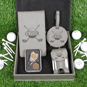 Puede incluir: Un juego de regalo de golf gris con artículos personalizados, que incluyen un bloc de notas, una etiqueta de equipaje y un marcador de bola. El juego incluye pelotas de golf y tees, todo dispuesto en una caja gris oscuro.