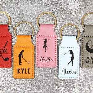 May include: Five personalized leather keychains with golf-themed designs. The keychains are in red, orange, pink, light blue, and gray. The designs include golf clubs, a golfer, and a golf ball. The keychains are personalized with the names "Mark", "Kyle", "Kristen", "Alexis", and "Coach Dallas".