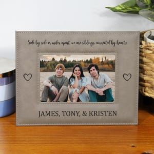 May include: A rectangular, light gray photo frame with a picture of three people. The frame has the text "Side by side or miles apart, we are siblings connected by heart" and the names "JAMES, TONY, & KRISTEN". Two heart symbols are on the frame.