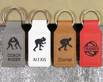 May include: Four leather keychains with silver key rings. The keychains are gray, white, brown, and red. Each keychain has a black silhouette of a wrestler and a name. The gray keychain says "COACH ROGER". The white keychain says "ALEXIS". The brown keychain says "DANIEL". The red keychain says "TANNER".