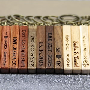 Puede incluir: Llaveros de madera con mensajes y diseños grabados. Los llaveros muestran frases como "Tyler", "Love, Jessica" y "Adventure Awaits". Cada llavero rectangular está unido a un anillo de metal.