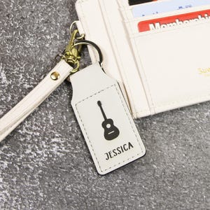 May include: A white leather keychain with a black guitar graphic and the name "JESSICA" printed below. The keychain is attached to a gold-colored metal ring and a matching wrist strap. A white wallet is in the background.