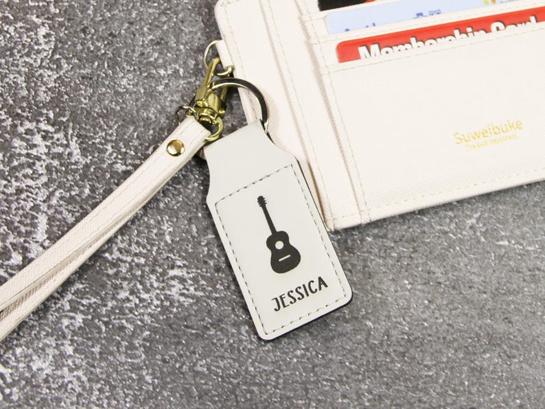 May include: A white leather keychain with a black guitar graphic and the name "JESSICA" printed below. The keychain is attached to a gold-colored metal ring and a matching wrist strap. A white wallet is in the background.