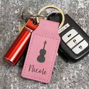 May include: A pink leather keychain with a black violin silhouette and the name "Nicole" printed on it. The keychain is attached to a set of keys, a red cylindrical object, and a car key fob.