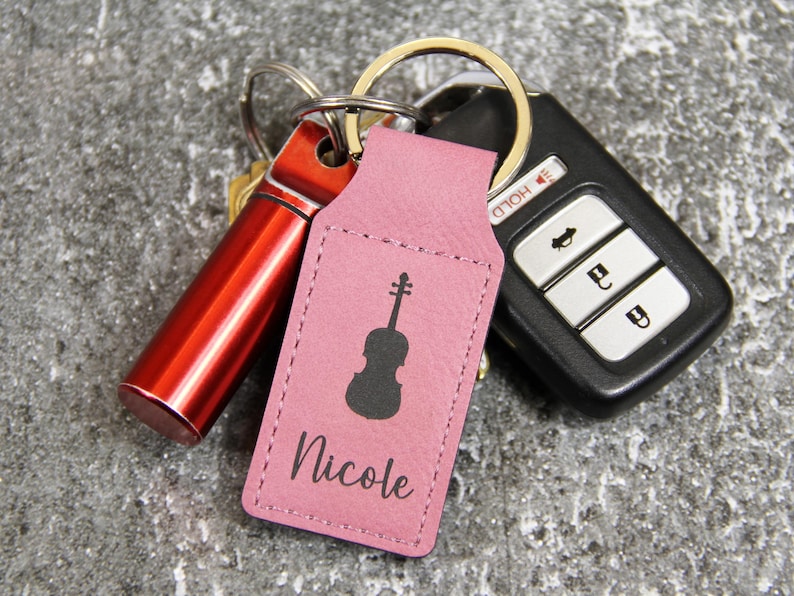 May include: A pink leather keychain with a black violin silhouette and the name "Nicole" printed on it. The keychain is attached to a set of keys, a red cylindrical object, and a car key fob.