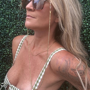 May include: A gold-colored eyeglass chain is draped over the chest. The chain is attached to a pair of sunglasses. The person is wearing a green and white gingham top. The background is a green hedge.