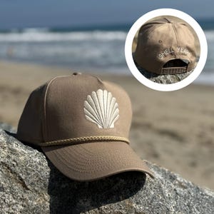 May include: A tan baseball cap with a white embroidered seashell design on the front. The cap has a rope detail along the brim and the text "SHELL YEAH!" embroidered on the back. The cap is sitting on a rock near the ocean.