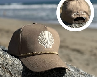 May include: A tan baseball cap featuring a white embroidered seashell design. The cap has a rope detail along the brim and the text "SHELL YEAH!" embroidered on the back. The cap is resting on a rock near the sea.