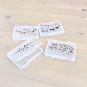 May include: Four rectangular, translucent plastic storage cases with hinged lids. Two cases feature black line art of beach-themed items and the text 'salty kiss swim'. One case displays a string of shells and swimwear, while another shows various sea creatures.