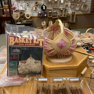 May include: A basket weaving kit for making an egg basket. The kit includes natural colored reeds and instructions. The kit is packaged in a clear plastic bag with a pink and white woven basket on display.