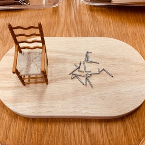 May include: A miniature wooden chair with a woven seat sits on a wooden board with a pile of silver metal hooks.