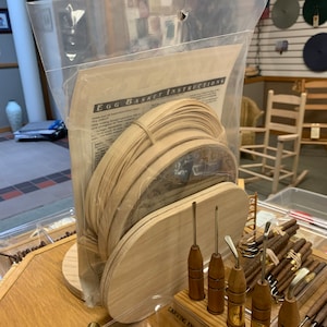 May include: A clear plastic bag containing a kit for making a woven egg basket. The kit includes wooden strips, a wooden base, and instructions. The bag is sitting on a wooden table with a variety of carving tools.