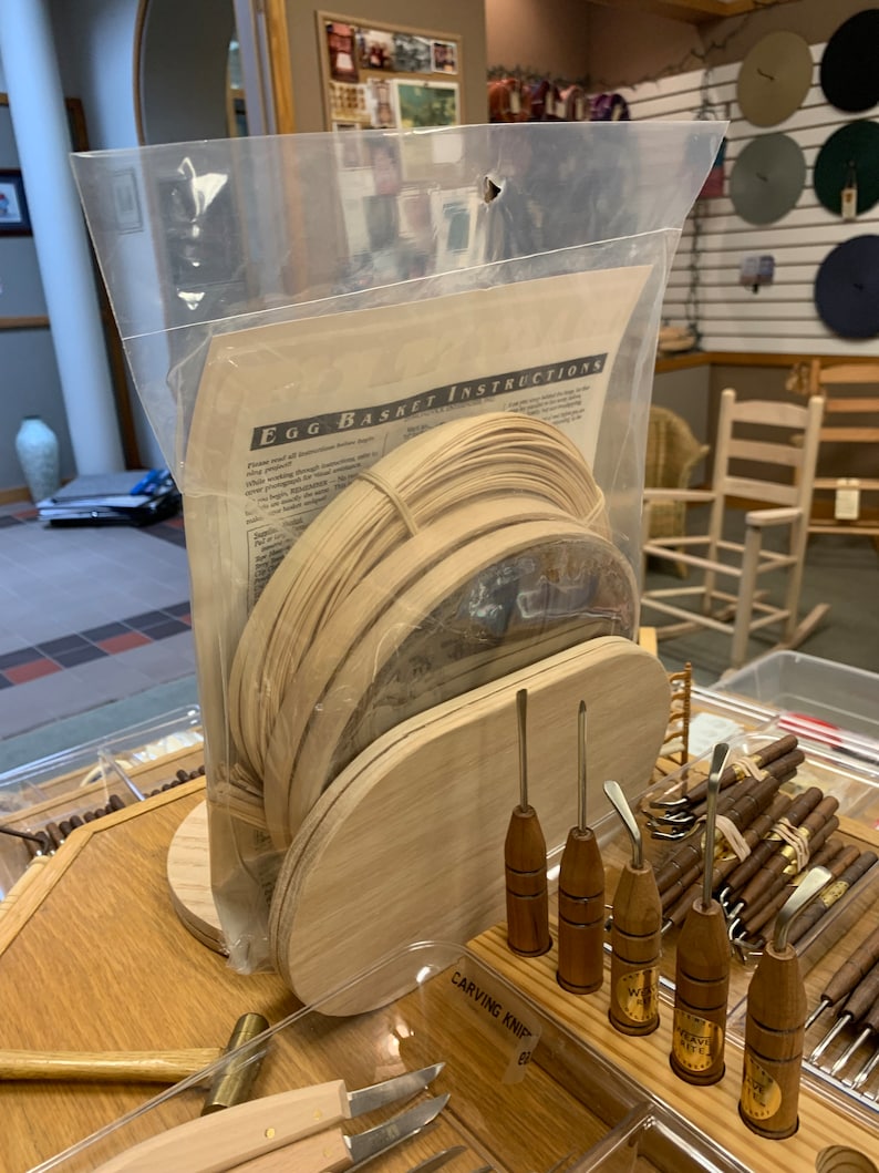 May include: A clear plastic bag containing a kit for making a woven egg basket. The kit includes wooden strips, a wooden base, and instructions. The bag is sitting on a wooden table with a variety of carving tools.