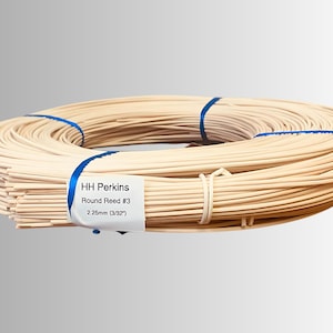 May include: A coiled bundle of round reed, secured with blue ribbon and a white label. The label reads "HH Perkins Round Reed #3 2.25mm (3/32")". The reed is a light beige color, suitable for crafts and basketry.