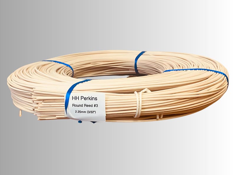 May include: A coiled bundle of round reed, secured with blue ribbon and a white label. The label reads "HH Perkins Round Reed #3 2.25mm (3/32")". The reed is a light beige color, suitable for crafts and basketry.