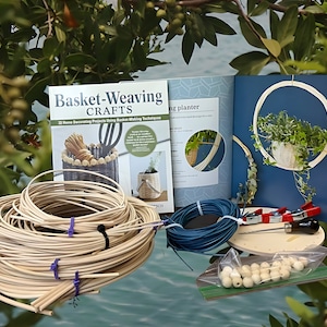 May include: A basket weaving crafts book with the title "Basket-Weaving Crafts: 22 Home Decorating Projects Using Basket Making Techniques". The book is open to a page showing a hanging planter made from woven reeds. The image also includes a bundle of natural colored wicker, blue twine, a wooden base, a set of wooden beads, and two red and black clamps.