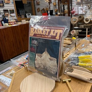 May include: A basket weaving kit for making an egg basket. The kit includes a wooden base, instructions, and reeds. The kit is packaged in a clear plastic bag with a red, white, and black label that says "Basket Kit Egg Basket #73022".