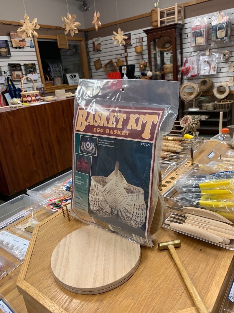 May include: A basket weaving kit for making an egg basket. The kit includes a wooden base, instructions, and reeds. The kit is packaged in a clear plastic bag with a red, white, and black label that says "Basket Kit Egg Basket #73022".