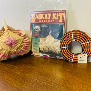 May include: A wicker egg basket kit with a finished basket, instructions, and a roll of 1/2 inch reed. The kit includes a pink and natural colored basket, a brown and orange roll of reed, and a printed instruction sheet with the title "Basket Kit Egg Basket".