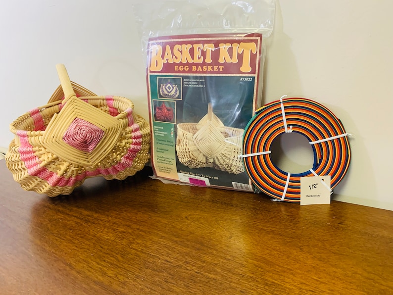 May include: A wicker egg basket kit with a finished basket, instructions, and a roll of 1/2 inch reed. The kit includes a pink and natural colored basket, a brown and orange roll of reed, and a printed instruction sheet with the title "Basket Kit Egg Basket".