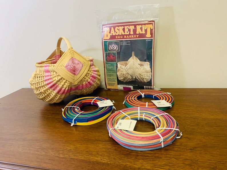 May include: A wicker egg basket kit with instructions and colorful reeds for weaving. The basket is pink and white with a diamond pattern. The reeds are in three bundles, each with a different color combination.