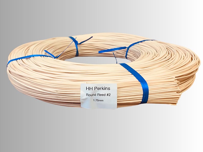 May include: A coiled bundle of light-colored round reed, secured with blue ribbon. A white label displays the text "HH Perkins Round Reed #2 1.75mm". This reed is likely used for weaving or crafting projects.
