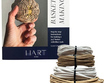 May include: A book titled "Basket Making" with a blue cover and the text "HART by hand" on the bottom. The book is next to a stack of three spools of brown and white fiber rush, which are used for basket weaving.