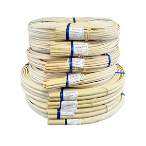May include: A stack of natural colored, round, woven cane, rattan, or wicker strips in various sizes. The strips are bundled together with blue twine. The labels on the bundles indicate the size of the strips in inches.