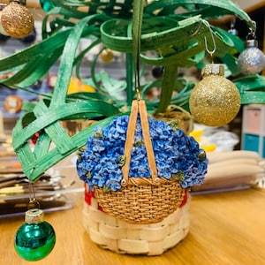 May include: A blue and green Christmas ornament shaped like a basket filled with blue flowers. The ornament is hanging from a green and brown tree.