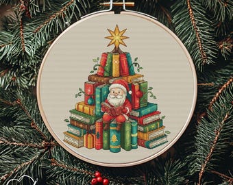 May include: A cross-stitch embroidery hoop featuring a Christmas tree made of books. Santa Claus is reading a book while sitting on the books. The tree is topped with a gold star. The background is a neutral color, and the hoop is surrounded by evergreen branches.