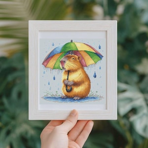 May include: A framed cross-stitch of a capybara sheltering under a rainbow umbrella. The capybara is a warm brown colour, and the umbrella has rainbow stripes. The background shows rain and grass, all within a white wooden frame.