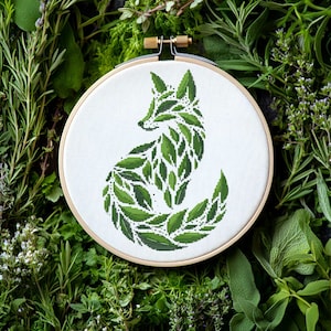 Puede incluir: Un aro de bordado blanco con un diseño de zorro verde hecho de hojas. El zorro está sentado con la cola enroscada alrededor de su cuerpo. El aro está rodeado de follaje verde.