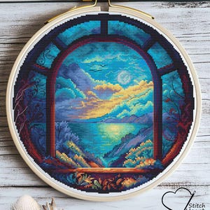 Puede incluir: Bordado de punto de cruz de un paisaje de atardecer visto a través de una ventana. La obra presenta un cielo nublado con montañas y un cuerpo de agua. Enmarcado en un bastidor de madera.
