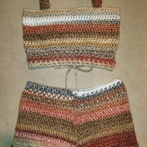 May include: A two-piece crochet set in earthy tones. The top is a crop top with straps, and the shorts have a drawstring waist. The set features horizontal stripes of brown, beige, white, and red.