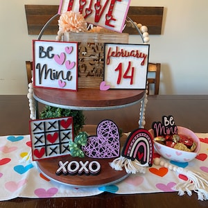 以下が含まれることがあります： バレンタインデーの装飾が施された2段の木製トレイ。上段には「LOVE」と「Be Mine」と書かれた看板があります。下段には三目並べゲーム、ハート、文字「XOXO」があります。チョコレートの小さなボウルも存在します。