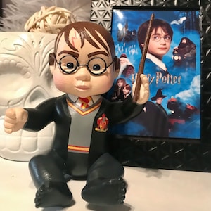 May include: A small, hand-painted figurine of Harry Potter as a baby, wearing a black robe with a Gryffindor crest and holding a wand. The figurine is sitting on a white surface in front of a framed poster of the first Harry Potter movie.