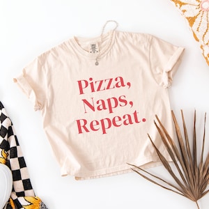 Könnte beinhalten: Beiges T-Shirt mit dem roten Aufdruck "Pizza, Naps, Repeat". Das Shirt liegt flach auf einer weißen Oberfläche, zusammen mit einer Halskette, einer gemusterten Decke, einem Kissen und einem getrockneten Palmblatt.